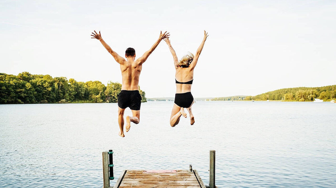 Ein Mann und eine Frau springen händehaltend in einen See