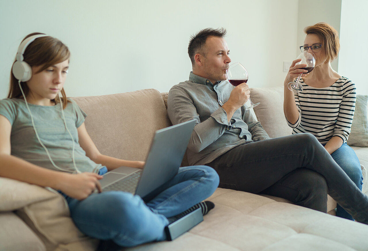 Eine Teenagerin sitzt mit Laptop auf dem Sofa neben zwei Erwachsenen, die Wein trinken